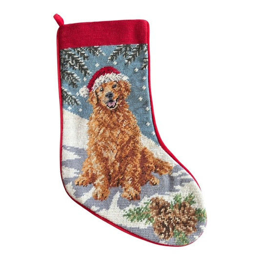 A Christmas stocking featuring a needlepoint-style design of a golden retriever dog wearing a Santa hat, set against a snowy winter background with pine trees and  pinecone. The stocking has a red border and a red hanging loop at the top.