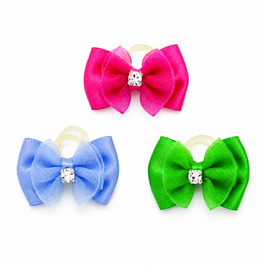 Three hair bows in bright pink, light blue, and green, each with a small clear rhinestone center and a beige elastic loop, shown on a white background.